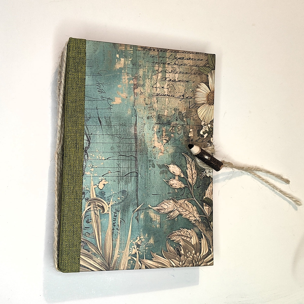 Foglie di Carta — quaderno tascabile fatto a mano con copertina botanica verde acqua, foglie e fiori su sfondo consumato come uno spartito antico, bordo in tessuto verde muschio, chiusura in spago naturale con bottone-matitina in legno
